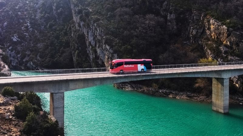 ônibus da blablacar passando por cima de uma ponte com montanhas e um rio ao fundo.